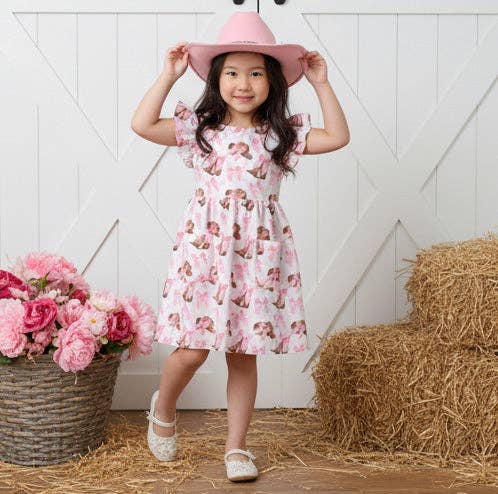 Young girl in a pink floral dress and hat standing on straw with flowers and hay bale in the background.