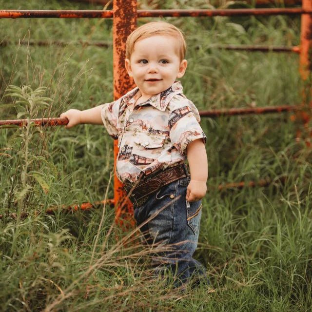 Child standing in a grassy field, wearing his train cowboy shirt next to a rusted fence