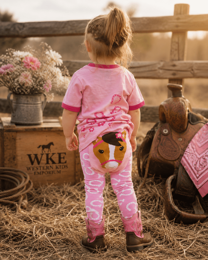 A toddler stands with their back to the camera in a rustic western-themed setting, wearing a pink short-sleeve t-shirt and matching pink leggings with white patterns and a cartoon horse graphic on the back.