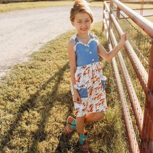 Child wearing western horse print dress with denim top, smiling and holding a fence.