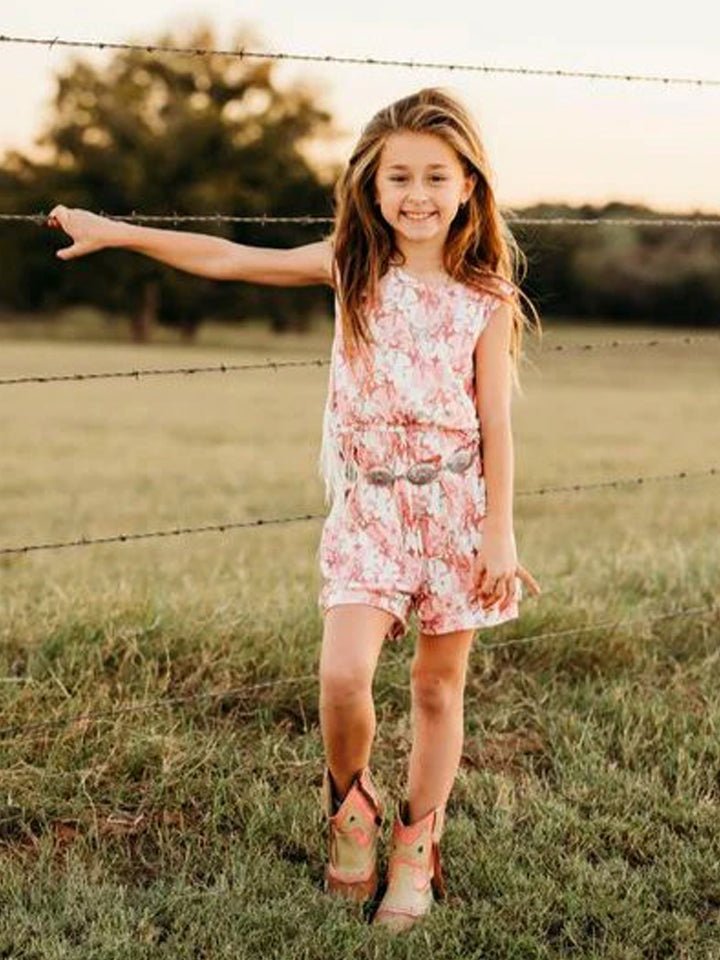 Smiling girl in pink plaid bucking horse fringe romper with cowboy boots, standing by a fence.