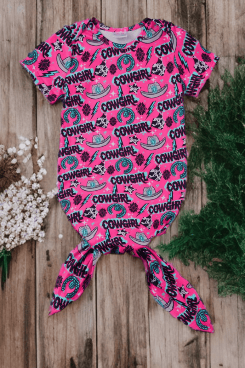 Pink cowgirl baby nightgown with Western prints and knot hem on wood background.