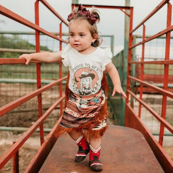 Toddler wearing Aztec western skirt beside red fence