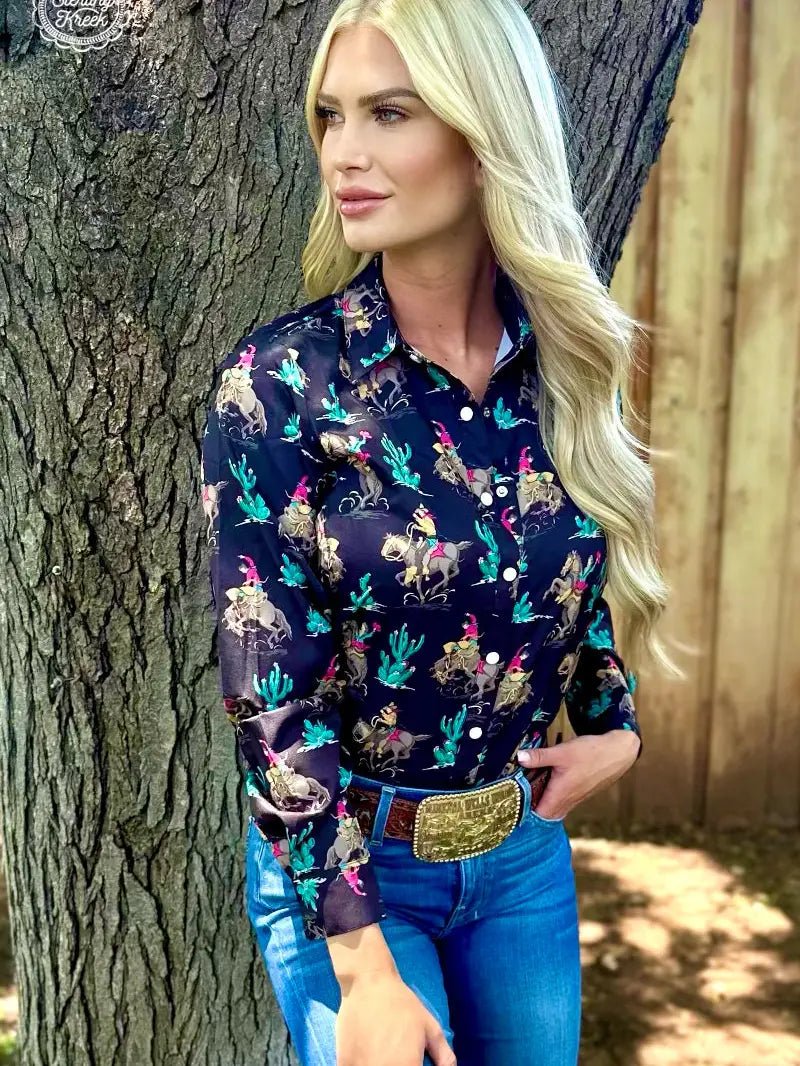 Blonde woman in black western grit shirt with rodeo print, standing by tree.