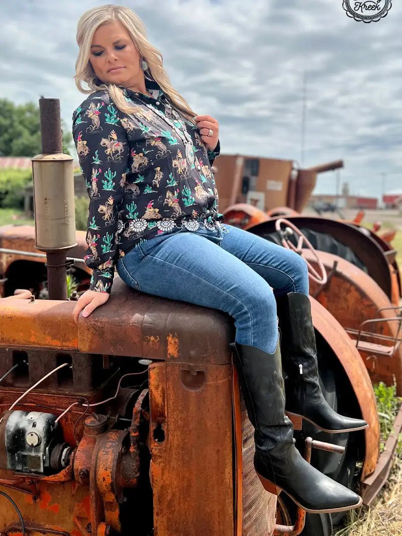 Woman in black western grit rodeo shirt sitting on old tractor, wearing jeans and boots.