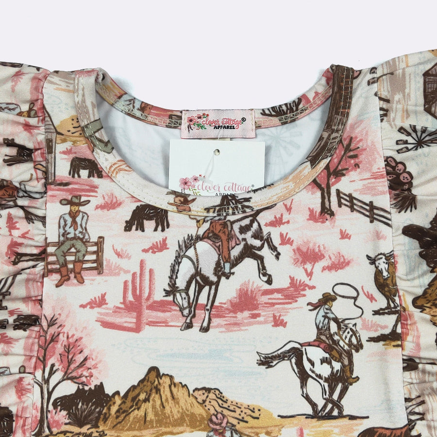 A close-up of a children's short-sleeve top featuring a western-themed print with cowboys, horses, cacti, and mountains in pink, brown, and beige tones. The inner neck label and hangtag show the brand 'The Cowgirl Collection'.