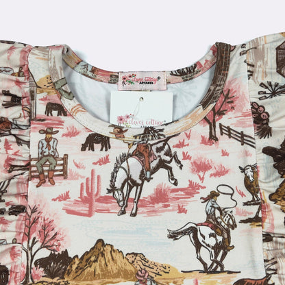 A close-up of a children's short-sleeve top featuring a western-themed print with cowboys, horses, cacti, and mountains in pink, brown, and beige tones. The inner neck label and hangtag show the brand 'The Cowgirl Collection'.
