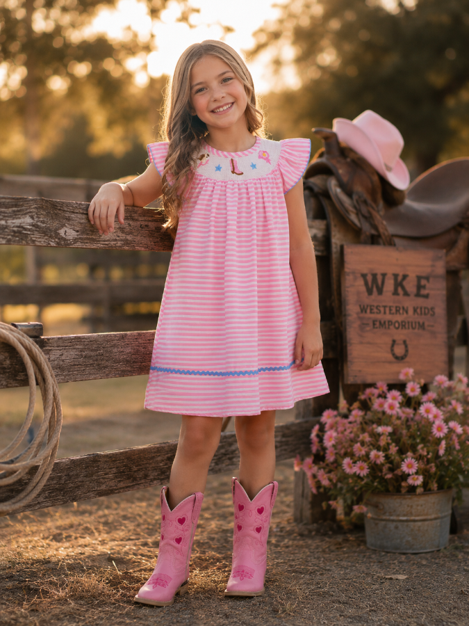 Young girl in a pink dress and cowboy boots standing next to a wooden fence with a cowboy hat and flowers in the background.