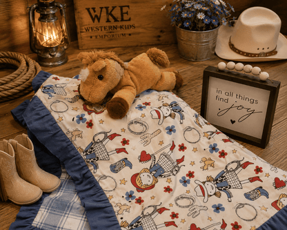 A western-themed baby blanket with a cowboy and cowgirl print, styled on a rustic wooden surface with a plush toy horse, cowboy boots, a lantern, a hat, and a decorative sign.