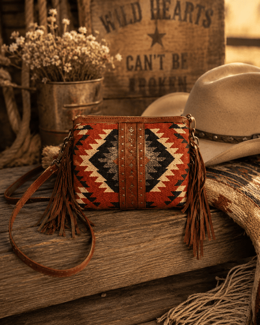 Woven handbag with tassels on a wooden surface with cowboy hat and flowers in the background.