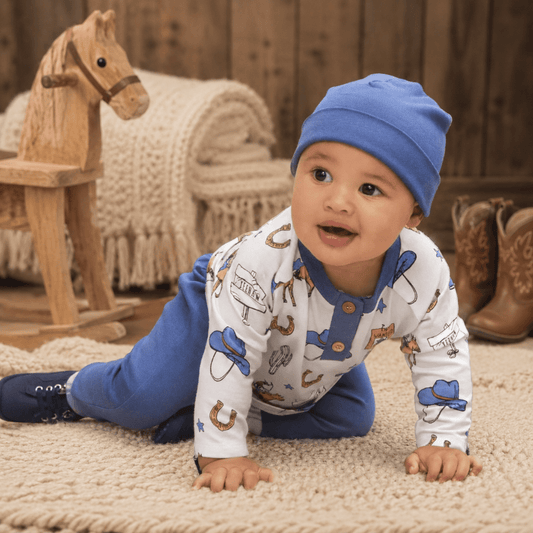 A baby crawling on a beige rug wearing a white long-sleeve onesie with a blue and brown cowboy print, blue pants, and a blue beanie hat. A wooden rocking horse and boots are in the background.