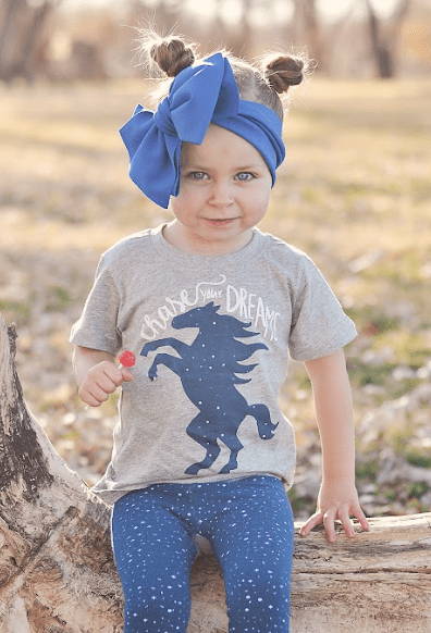 Child wearing a gray shirt with a horse graphic and blue leggings, standing outdoors.