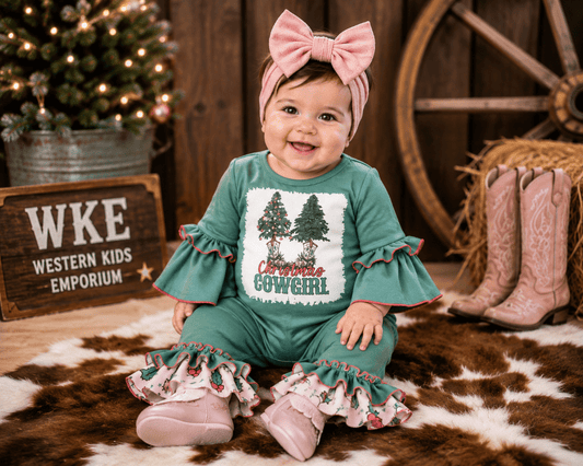 A baby girl wearing a green Christmas-themed ruffled one-piece outfit with a 'Christmas Cowgirl' graphic, a pink bow headband, and pink boots, sitting on a cowhide rug in a rustic holiday setting.