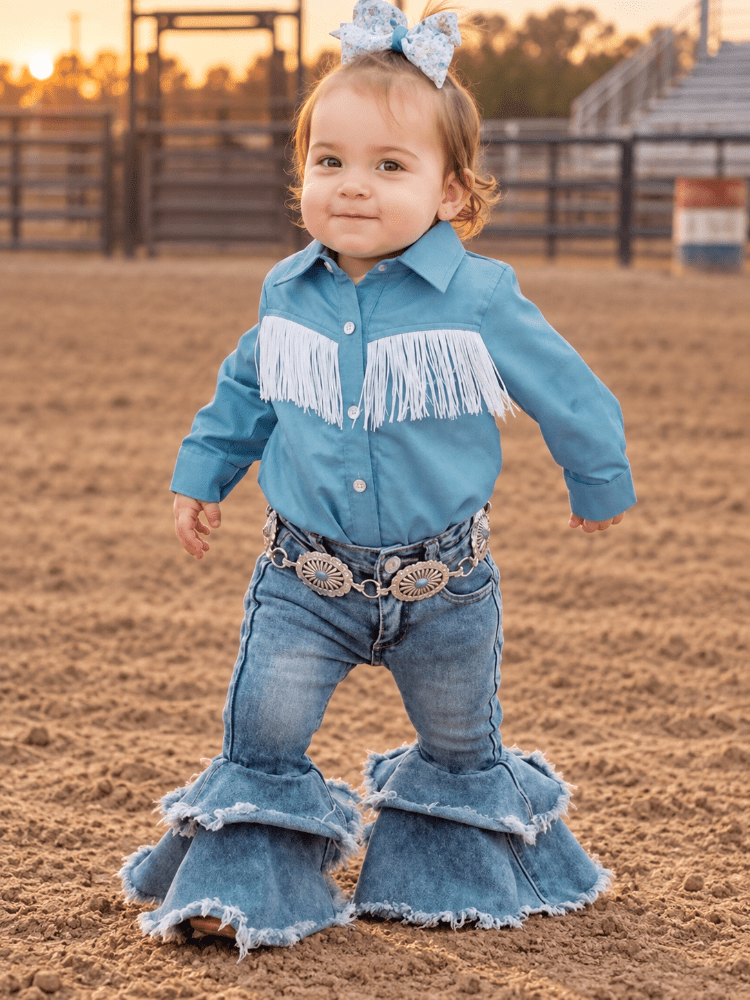 A toddler stands in a sandy outdoor arena wearing a western-style outfit with a light blue long-sleeve shirt featuring white fringe across the chest, matching light blue denim jeans with large ruffled hems, a decorative silver-toned belt, and a blue bow in her hair.