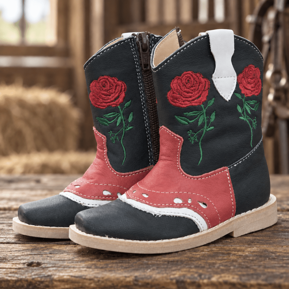 A pair of western-style boots with a black leather upper, red leather panels, green embroidered roses, and white pull tabs, displayed on a rustic wooden surface.