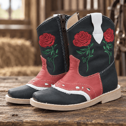 A pair of western-style boots with a black leather upper, red leather panels, green embroidered roses, and white pull tabs, displayed on a rustic wooden surface.