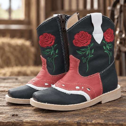 A pair of western-style boots with a black leather upper, red leather panels, green embroidered roses, and white pull tabs, displayed on a rustic wooden surface.