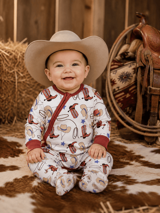 A smiling baby wearing a cowboy hat and a white footed sleeper with a western-themed print of boots, horseshoes, stars, and horseshoe patterns, sitting on a cowhide rug with a rustic western backdrop.