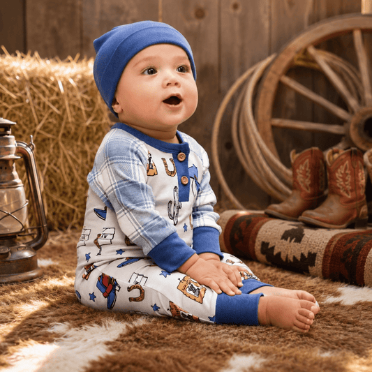 A baby wearing a blue knit beanie and a western-themed long-sleeve romper sits on a brown and white cowhide rug in a rustic western-themed setting with hay bales, a lantern, cowboy boots, and a wagon wheel.