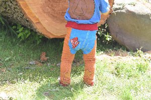 Baby wearing a blue shirt and chap leggings standing next to a large log in a grassy area.