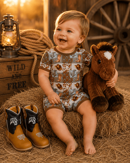 Child in a cowboy romper with boots and a horse plush toy, sitting on hay with a rustic background.