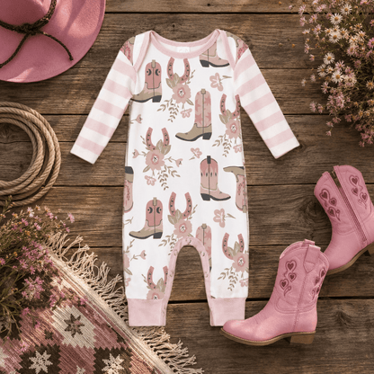 A baby long-sleeve footless romper laid flat on a rustic wooden surface, styled with pink cowboy boots, a pink hat, flowers, rope, and a patterned rug. The romper is white with light pink horizontal stripes on the sleeves and legs, and an allover print of pink and tan cowboy boots with floral details.
