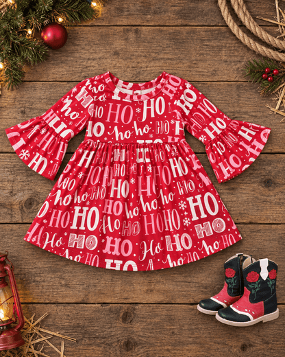 A festive red children's dress with a white 'HO HO HO' holiday print, styled in a flat-lay on a rustic wooden background with Christmas decorations and red cowboy boots.