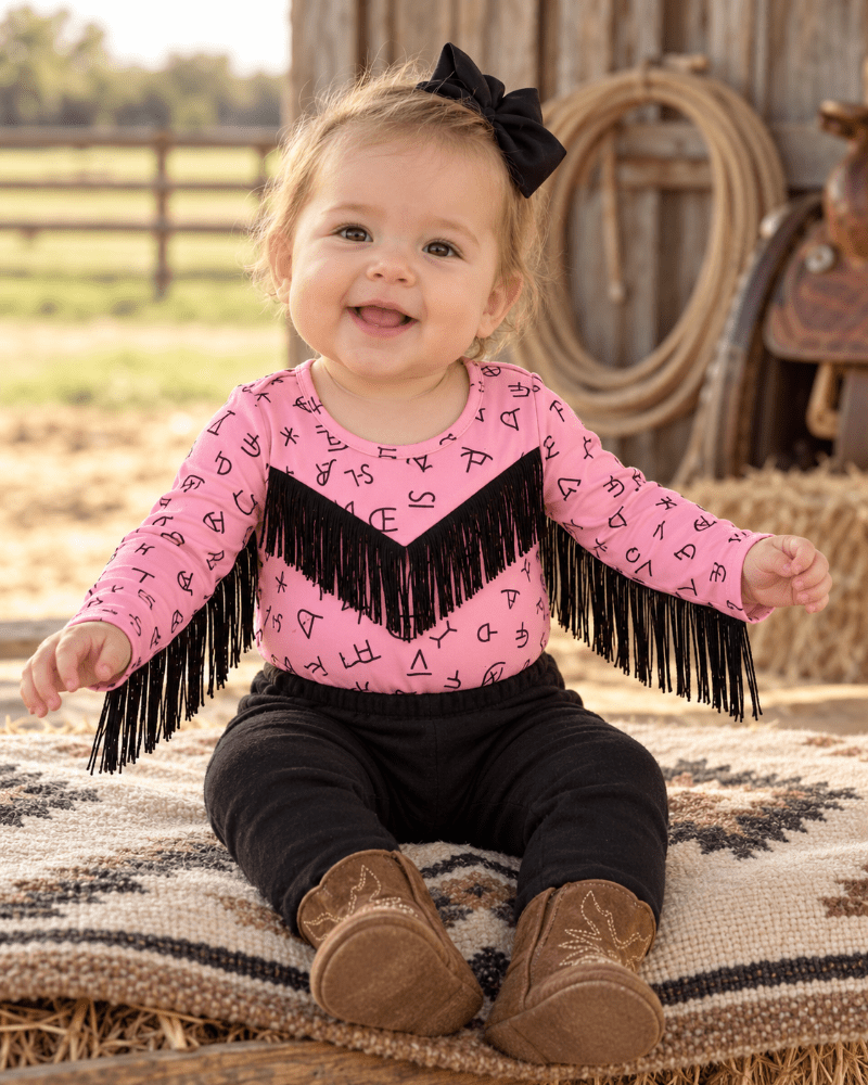 A baby girl sitting on a western-style blanket in a barn setting, wearing a pink long-sleeve top with black chevron fringe and letter print, black pants, and brown cowboy boots.