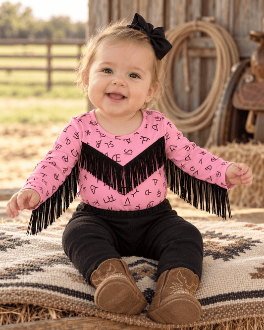 A baby girl sitting on a western-style blanket in a barn setting, wearing a pink long-sleeve top with black chevron fringe and letter print, black pants, and brown cowboy boots.