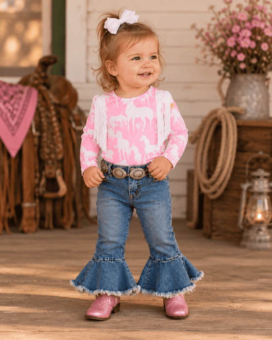 A toddler girl stands on a rustic porch wearing a pink long-sleeve shirt with white fringe and a pink-and-white animal print, blue flared jeans with a frayed hem, a western-style belt with large silver conchos, and pink cowboy boots.