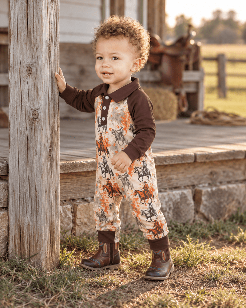 A toddler stands on a rustic porch wearing a brown long-sleeve collared jumpsuit with an orange, black, and beige western horse pattern, paired with dark brown boots.
