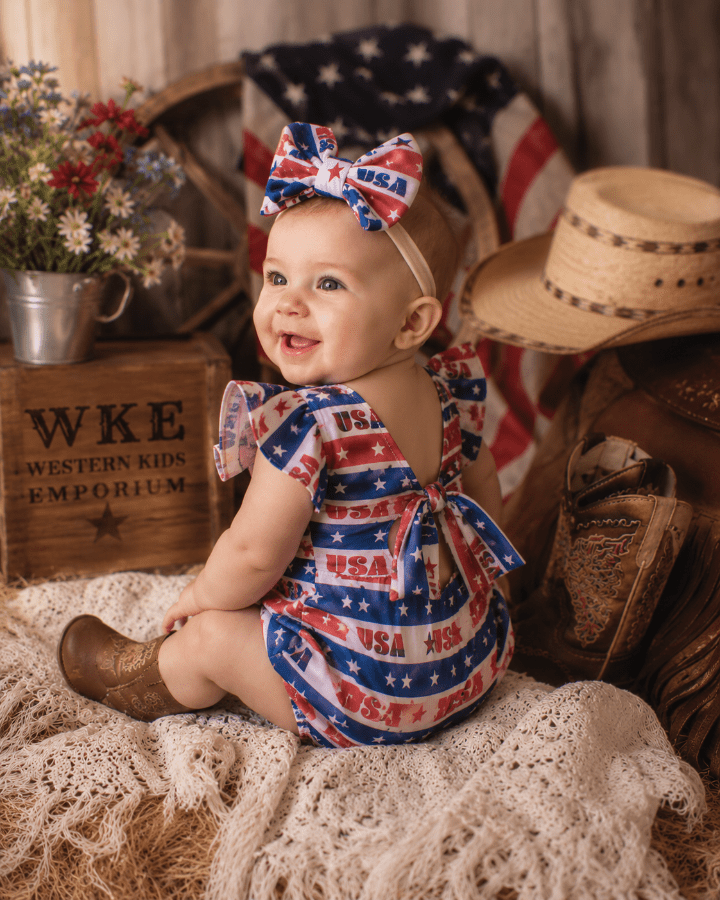 Baby in a patriotic outfit with an American flag headband, surrounded by rustic decor.