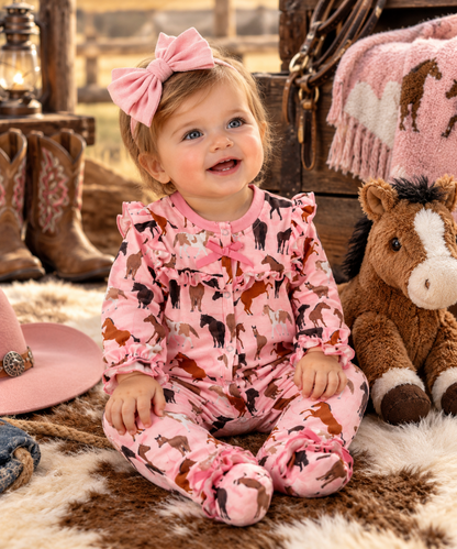 A baby girl wearing a pink long-sleeve footed jumpsuit with a cow and horse print, sitting on a cowhide rug in a rustic western-themed room. She has a matching pink bow in her hair.