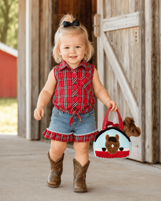 A toddler girl stands in front of a rustic wooden barn door, wearing a red plaid sleeveless button-up top, denim shorts with a plaid ruffle, and brown cowboy boots. She is holding a small red and white felt purse with a cartoon horse design.