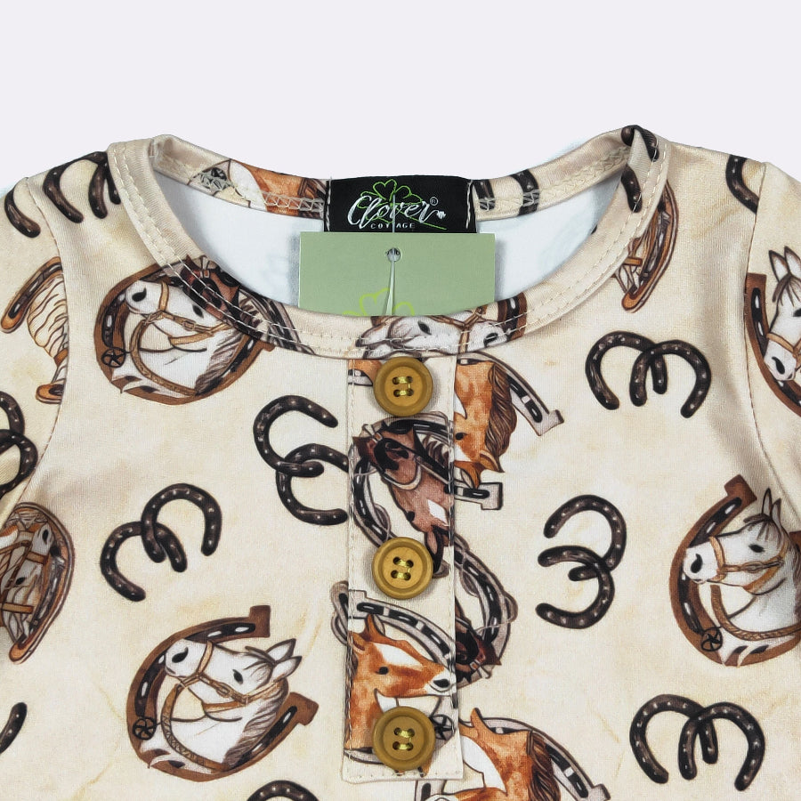A close-up of a baby long-sleeve top by Clover, featuring a cream-colored fabric with a western-themed print of horses, horseshoes, and a brown horse illustration. The top has a three-button placket with brown buttons and a light green brand tag still attached at the collar.