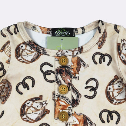 A close-up of a baby long-sleeve top by Clover, featuring a cream-colored fabric with a western-themed print of horses, horseshoes, and a brown horse illustration. The top has a three-button placket with brown buttons and a light green brand tag still attached at the collar.