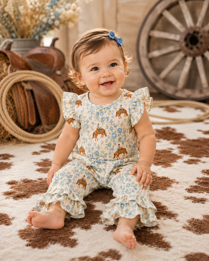 A smiling baby girl sitting on a cow-print rug, wearing a light blue and white floral romper with brown horse prints and ruffled sleeves and legs. She has a blue bow in her hair, with rustic western decor in the background.