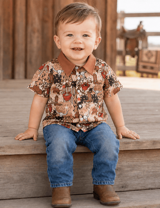 A smiling toddler boy sitting on wooden steps wearing a brown short-sleeve western-style button-up shirt with a cow print, blue jeans, and brown boots.