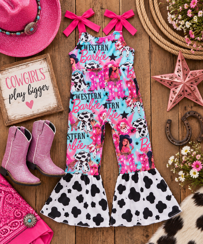 A children's sleeveless Western-themed jumpsuit with pink and turquoise cow-print panels, Dalmatian graphics, and black-and-white cow-print flared legs, styled on a wooden surface with pink cowboy boots, a pink hat, a sign reading 'Cowgirls play bigger,' and western decor.