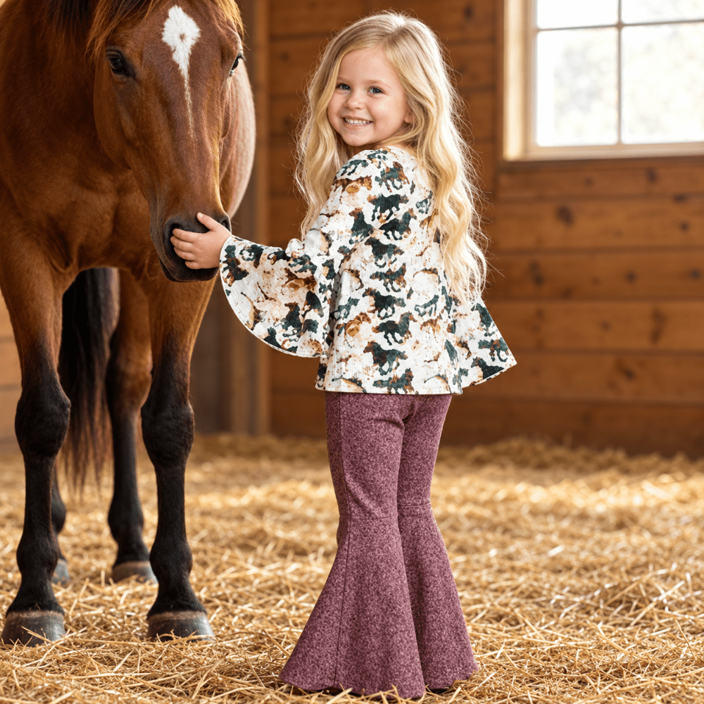 A young blonde girl stands in a barn next to a brown horse, wearing a white long-sleeve blouse with a green, brown, and tan animal print, paired with textured mauve-brown flared pants.
