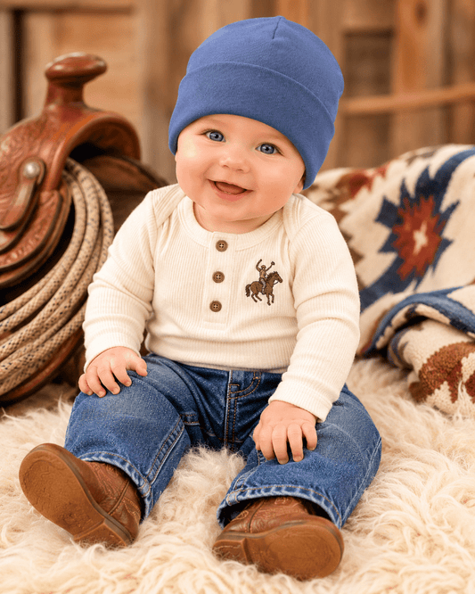 A baby sitting on a white faux fur rug wearing a cream ribbed henley shirt with a brown embroidered cowboy motif, blue jeans, and brown boots, while wearing a blue beanie. A western saddle and patterned blanket are visible in the background.