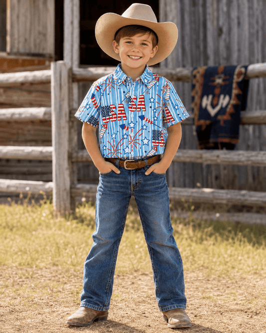 A young boy stands outdoors in front of a rustic wooden fence and barn, wearing a cowboy hat, a short-sleeve button-up shirt with a red, white, and blue American flag and fireworks print, blue jeans, and brown boots.