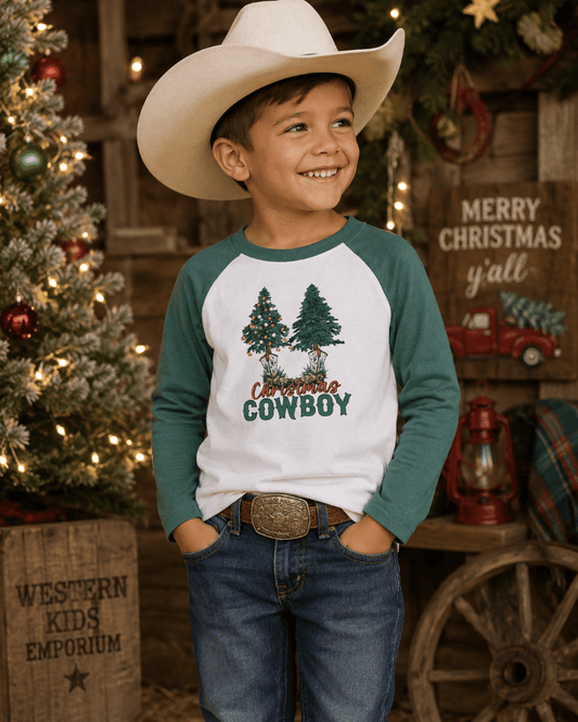 A young boy wearing a cream cowboy hat and a green and white raglan long-sleeve shirt with a 'Christmas Cowboy' tree graphic, standing in a rustic Christmas-themed store setting.