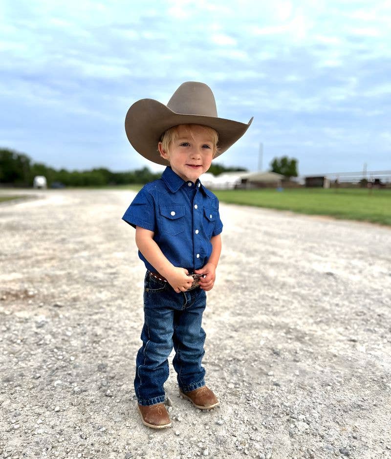 Child wearing a cowboy hat, blue shirt, and jeans standing on a gravel road with a clear sky.