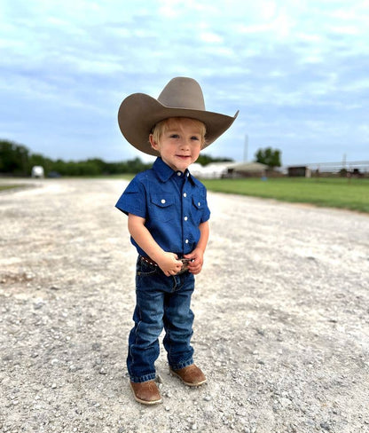 Child wearing a cowboy hat, blue shirt, and jeans standing on a gravel road with a clear sky.
