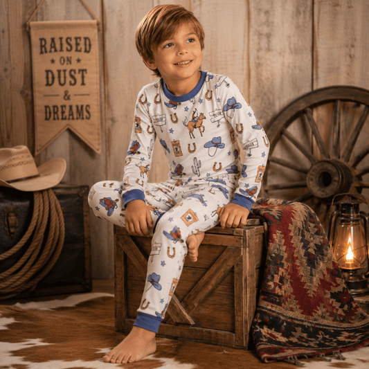 A young boy wearing a white long-sleeve pajama set with a cowboy-themed print, sitting on a wooden crate in a rustic western-themed room.