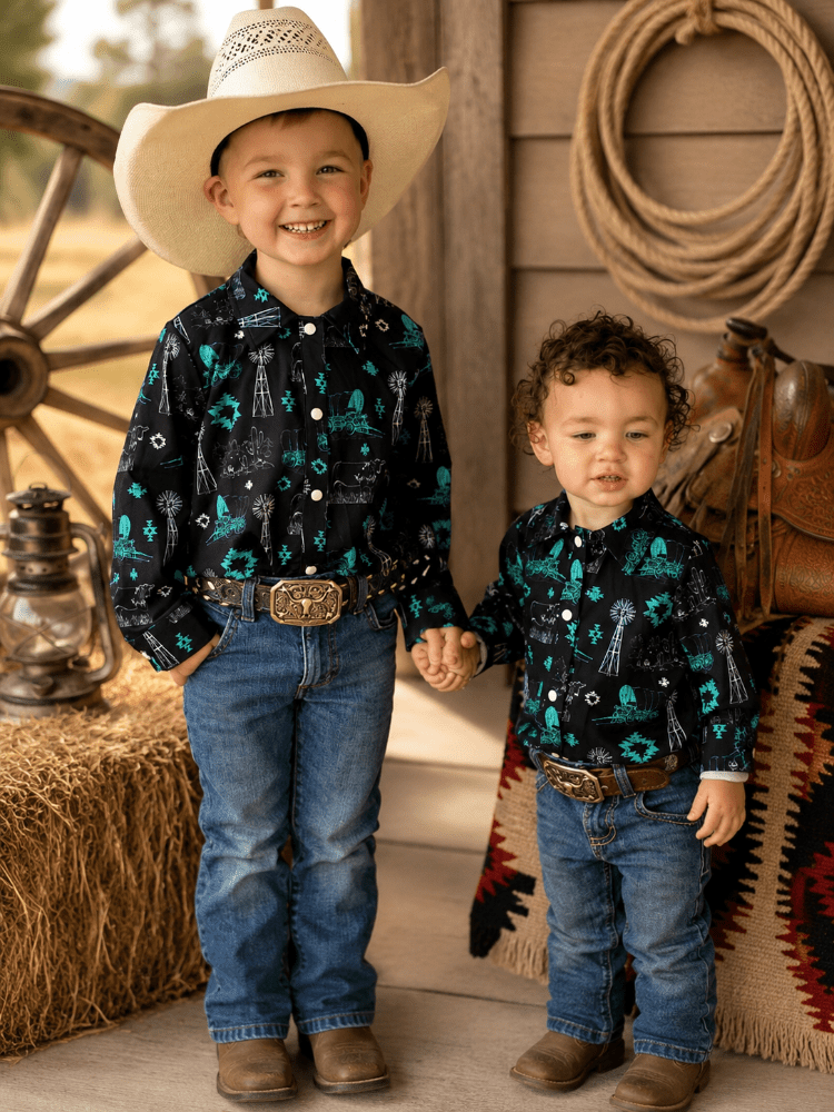 Two young boys standing together in a rustic western-themed setting. The boy on the left wears a large cream cowboy hat, a black long-sleeve button-up shirt with a teal and white skeleton/rusty pattern, blue jeans, and brown boots.