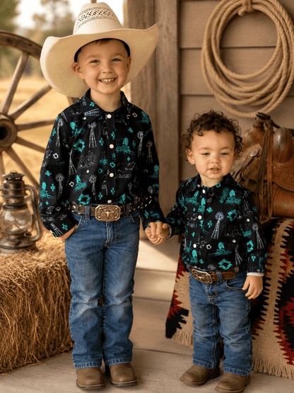 Two young boys standing together in a rustic western-themed setting. The boy on the left wears a large cream cowboy hat, a black long-sleeve button-up shirt with a teal and white skeleton/rusty pattern, blue jeans, and brown boots.