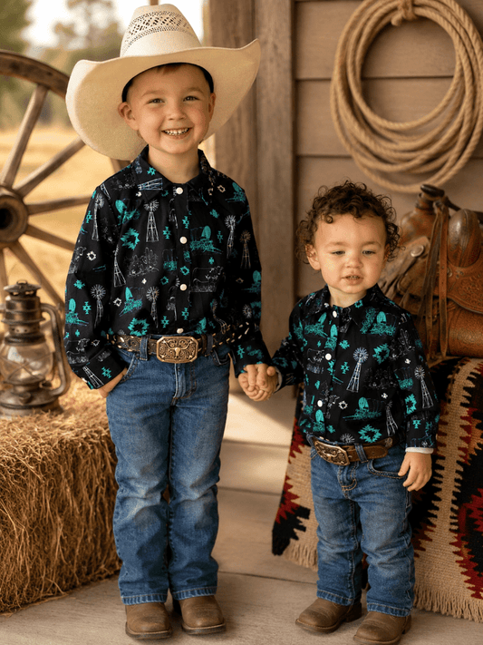 Two young boys standing together in a rustic western-themed setting. The boy on the left wears a large cream cowboy hat, a black long-sleeve button-up shirt with a teal and white skeleton/rusty pattern, blue jeans, and brown boots.