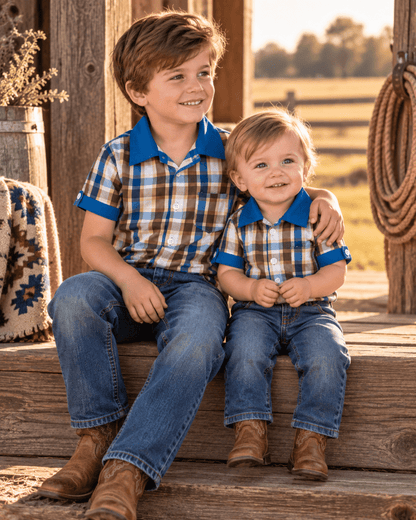Two children sitting together on rustic wooden steps outdoors, wearing matching plaid short-sleeve polo shirts with blue collars and cuffs, blue jeans, and brown boots.